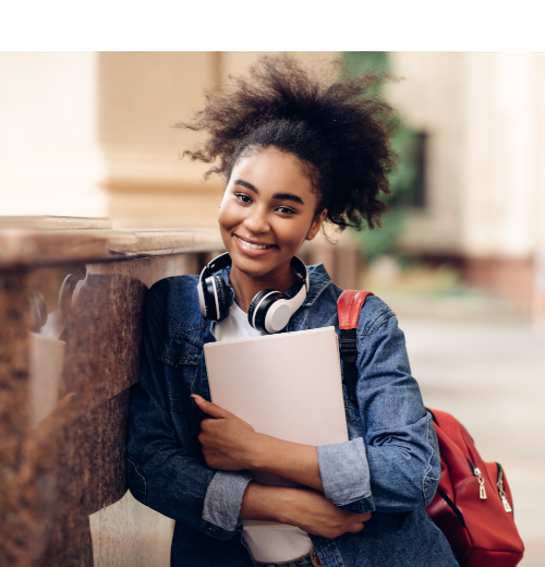 Smiling student standing outdoors, holding a notebook, wearing headphones around their neck and a backpack.