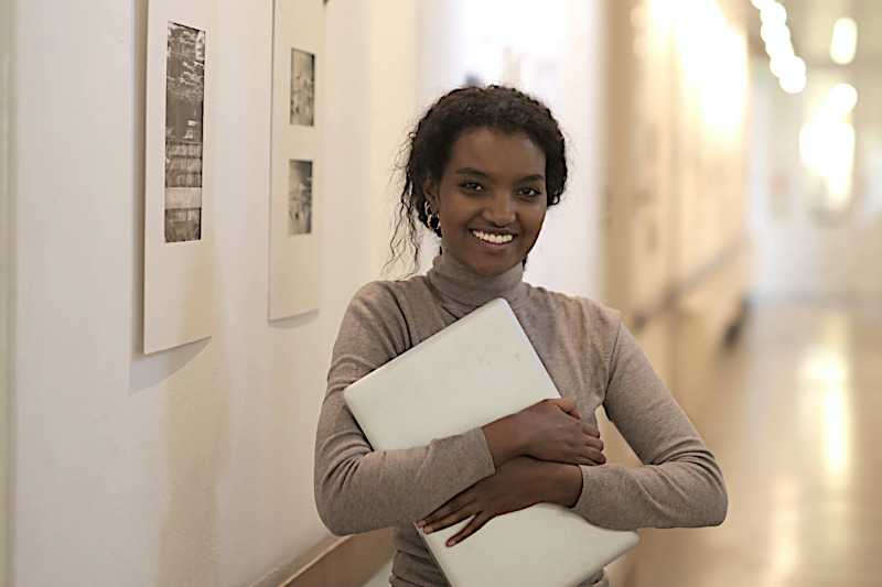 A young woman is holding her closed laptop and smiling into the camera.