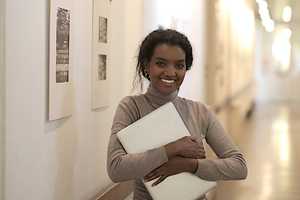 A young woman is holding her closed laptop and smiling into the camera.