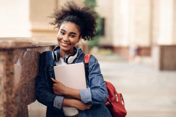 A student with curly hair leans against a marble surface, holding a laptop and wearing headphones, carrying a red backpack.