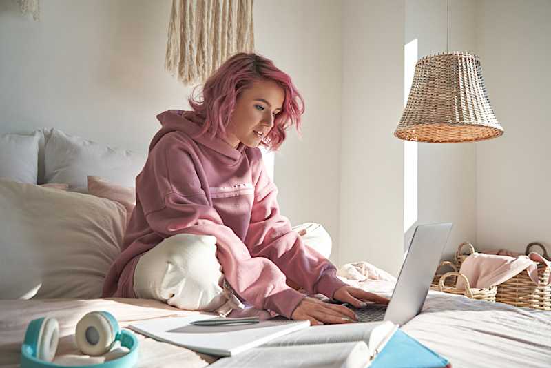 A young woman is in bed working at a laptop. 