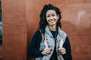 Smiling woman with curly hair stands against a textured, reddish-brown wall. She wears a gray vest and dark sweater, exuding warmth and confidence.