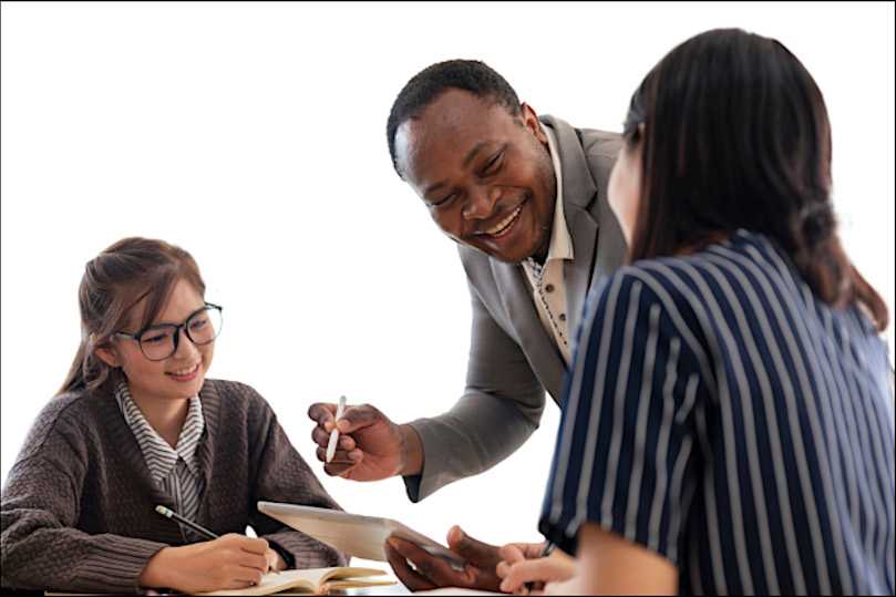 An adult educator smiles while pointing to a tablet as a student takes notes, with another adult reviewing information beside them.