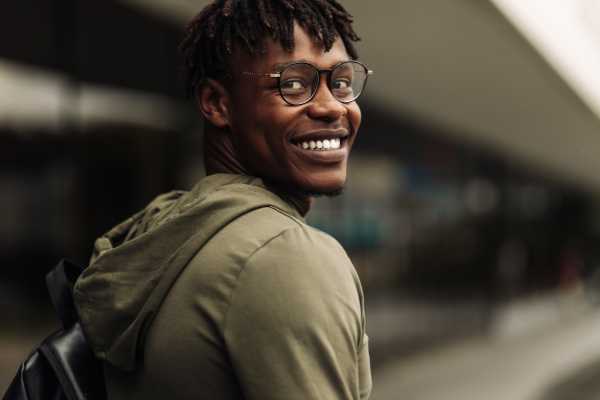 A young Black man with a backpack on is looking over he shoulder at the camera and smiling. 