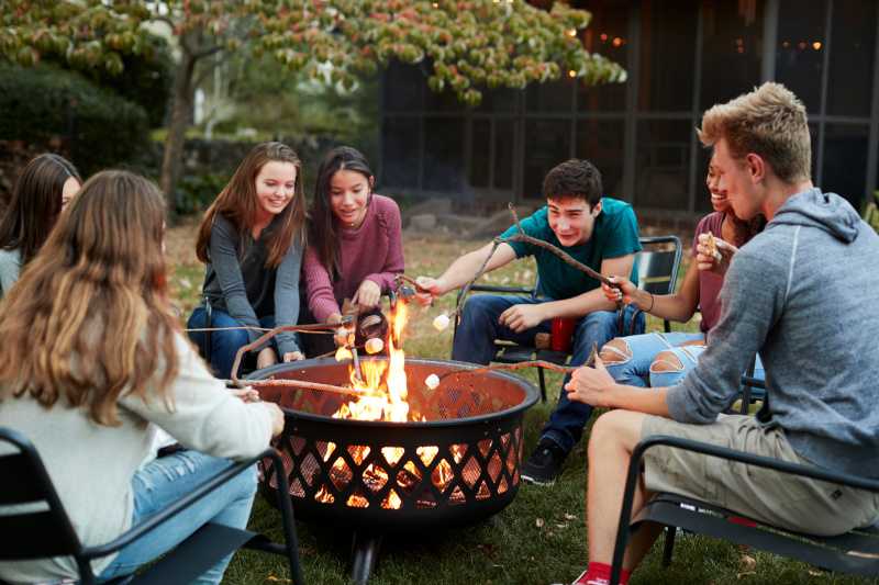 A group of young people are roasitng marshmallows around a campfire. 