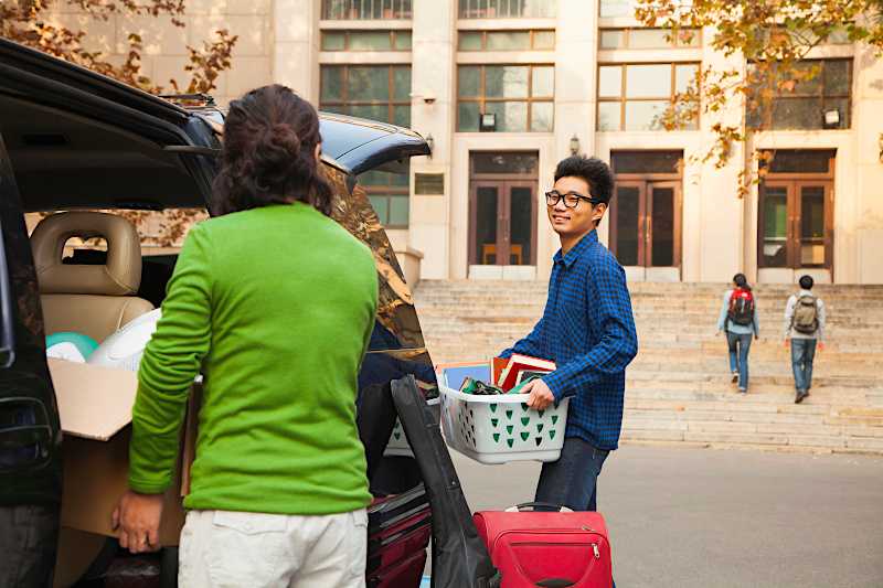 A mother and son are taking belongings out of the back of a car to move into the college dorm. 